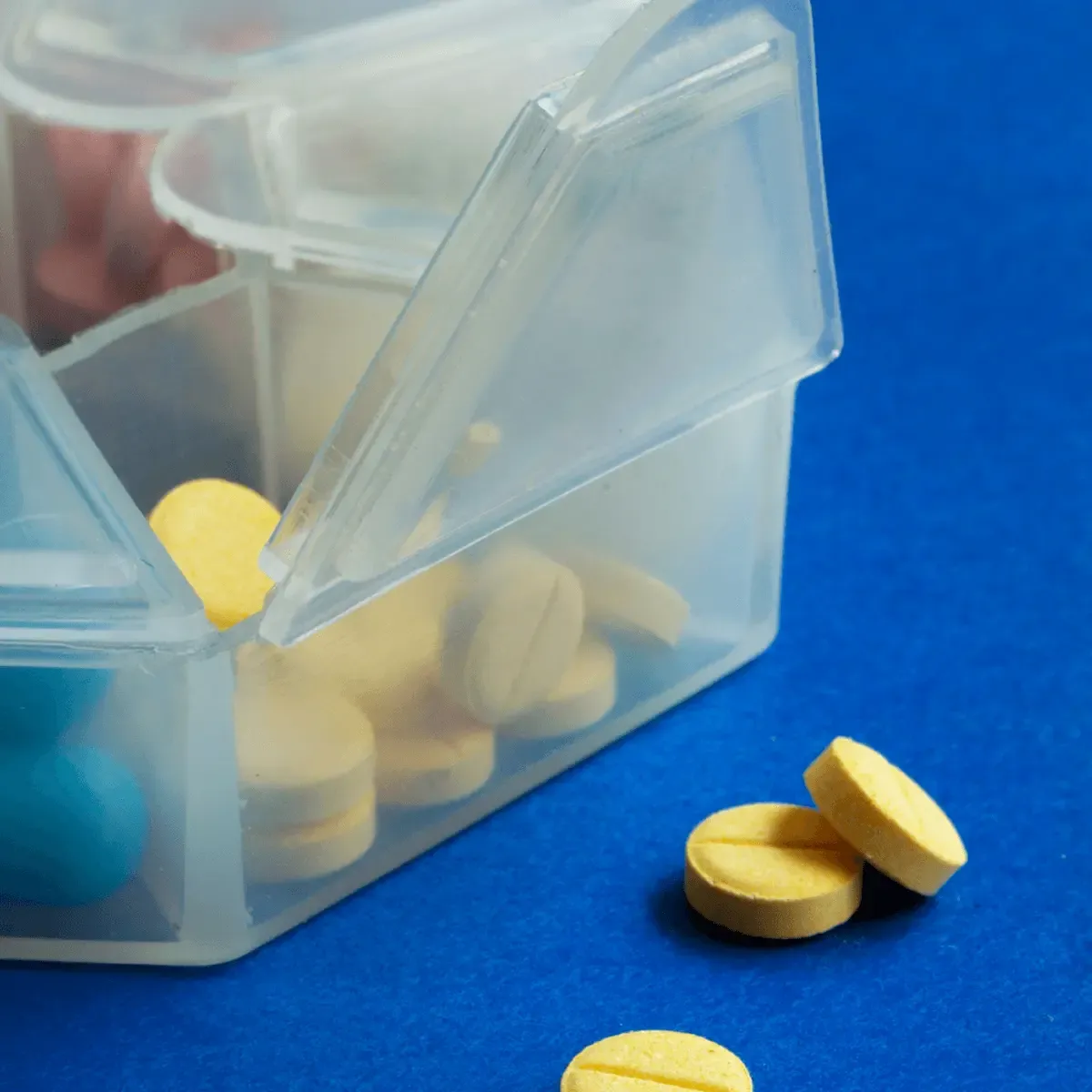 Clear plastic pill organizer container tipped over with yellow round tablets spilling out onto blue surface